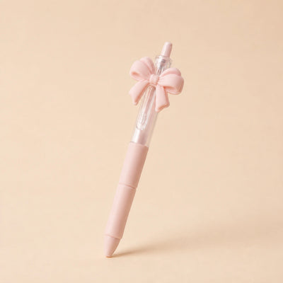 A pastel pink retractable pen with a decorative pink bow on the cap, standing upright on a solid beige background.