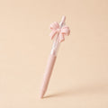 A pastel pink retractable pen with a decorative pink bow on the cap, standing upright on a solid beige background.