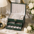 An open dark green jewelry organizer box with two drawers, filled with silver and pearl jewelry, styled on a table with flowers, a lamp, and a small bottle.