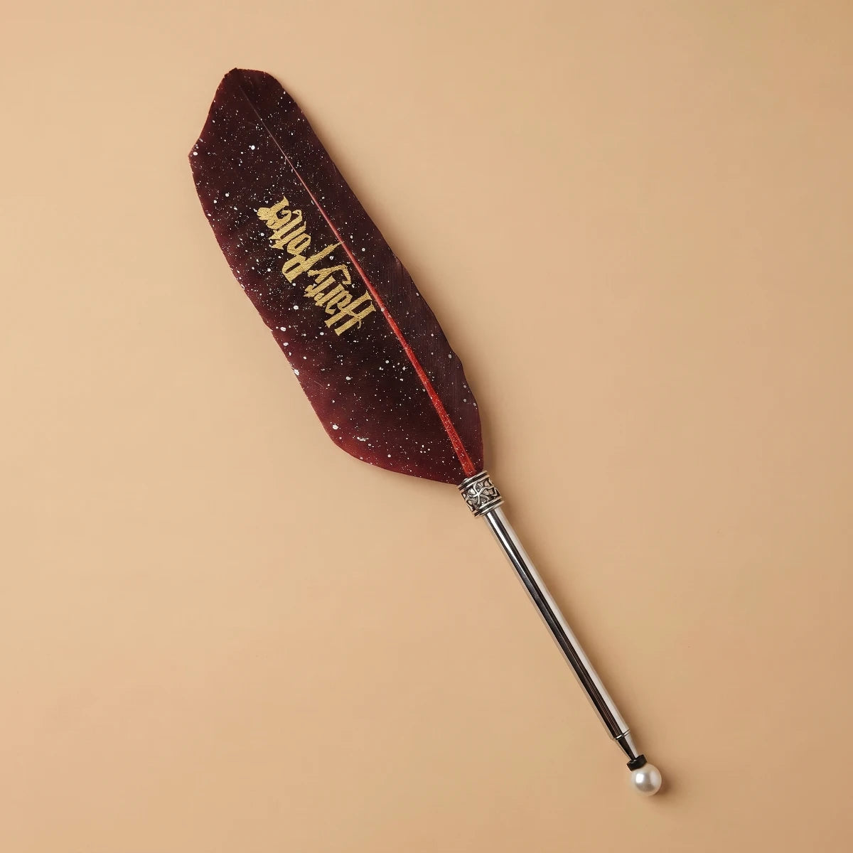 A Harry Potter themed feather quill accessory with a dark burgundy glittery feather printed with gold 'Harry Potter' text, attached to a silver metal rod with a pearl finial, set against a solid beige background.