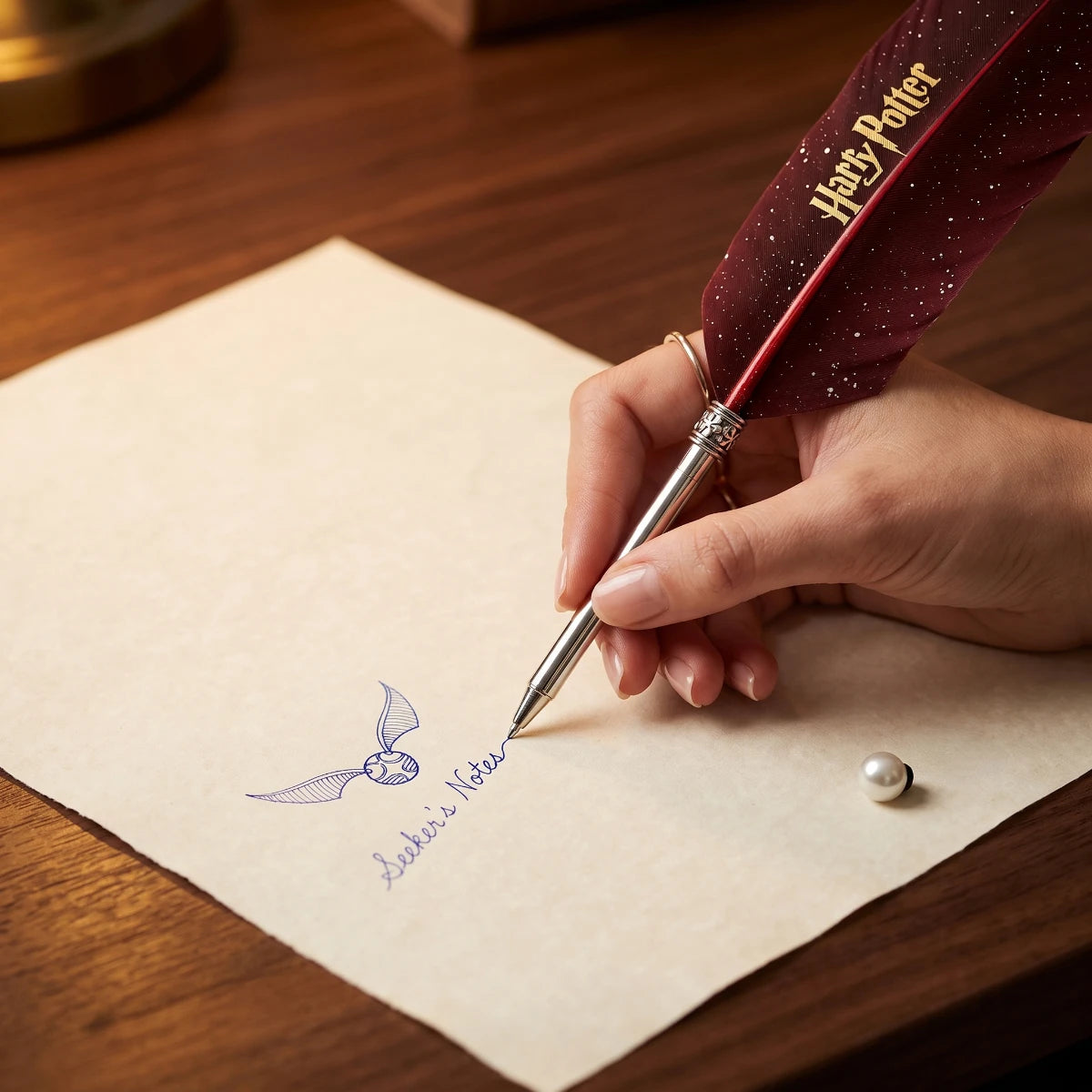 A hand holds a decorative quill pen with a dark red feather and gold 'Harry Potter' text, writing on cream paper over a wooden desk. A small pearl-like bead rests nearby.