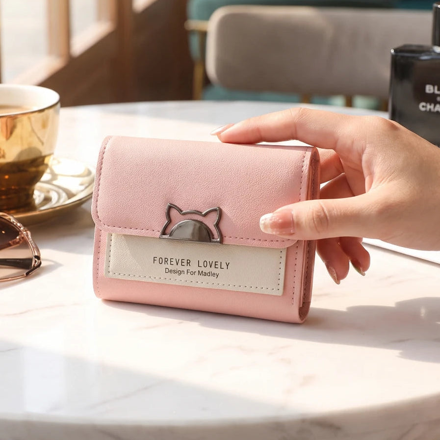A hand holds a light pink bifold wallet with a cat face charm and a white label reading 'FOREVER LOVELY Design For Millennials' on a marble table. A gold coffee cup, sunglasses, and a Chanel perfume bottle are nearby in a cafe setting.