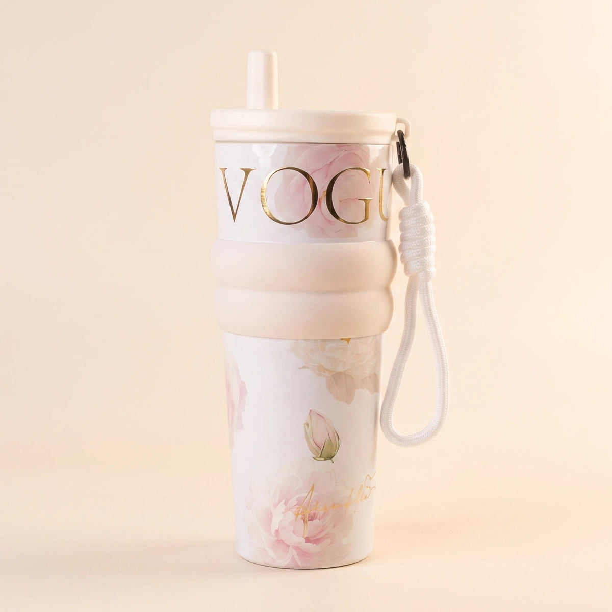A tall, slim travel tumbler with a white lid and straw, featuring a pink floral print, a beige silicone sleeve with gold 'VOGUE' lettering, and a white wrist strap, set against a warm beige background.