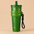 A green St. Patrick's Day themed tumbler with a shamrock pattern, featuring the brand name 'OPINIA' in gold lettering, a matching green straw lid, a green silicone band, and a black wrist strap, set against a solid beige background.