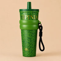 A green St. Patrick's Day themed tumbler with a shamrock pattern, featuring the brand name 'OPINIA' in gold lettering, a matching green straw lid, a green silicone band, and a black wrist strap, set against a solid beige background.